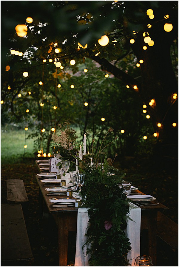 An outdoor evening reception tablescape set in a wooded garden, photographed as a wide detail shot with no guests present. A long rustic wooden farm table with bench seating is dressed with a white linen runner layered with a dense greenery garland featuring ferns and dark foliage. White taper candles and small votive candles are placed along the centerpiece, alongside crystal glassware, white plates, and what appears to be gold-accented tableware. String globe lights are strung through the tree branches overhead, creating warm amber lighting in the dim evening setting. The overall styling is rustic-botanical with a dark, moody atmosphere.