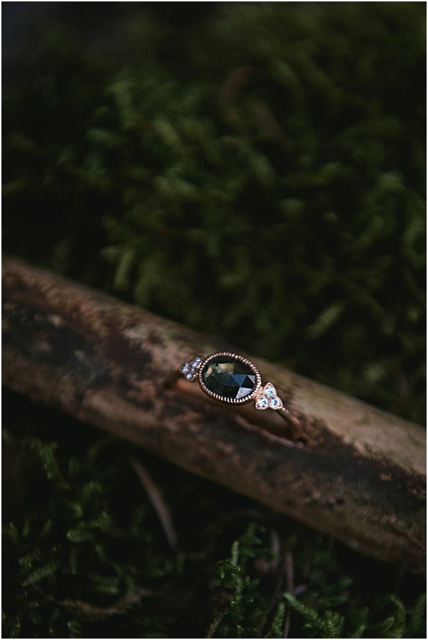A close-up detail shot of an engagement or wedding ring resting on a small weathered wood branch surrounded by dark green moss. The ring features a rose gold band with a milgrain or beaded texture, a large oval dark blue-green or black rose-cut center stone, and small diamond accent clusters on either side of the setting. The shallow depth of field keeps the ring in sharp focus while the mossy background blurs softly, creating a nature-inspired, moody composition with dark, muted tones throughout.