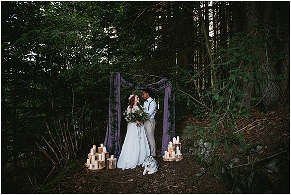 A couple stands together in a forest clearing beneath a wooden arch draped with dusty purple/mauve fabric, facing each other and smiling. The bride wears a flowing white gown and holds a bouquet of greenery and white blooms, while the groom wears light grey trousers with suspenders and a white shirt. A husky dog sits at their feet between groupings of pillar candles arranged on raw wood slice platforms on either side of the arch. The styling is boho-rustic with a muted purple and natural wood palette, set entirely outdoors among tall dense trees in low ambient light. The image is a medium-wide portrait shot capturing the full ceremony setup.