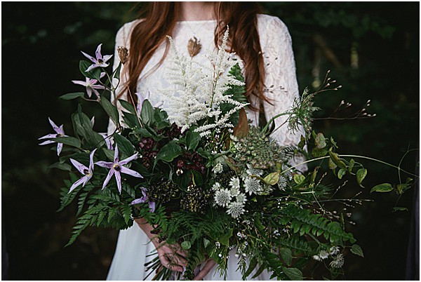 Close-up portrait of a bride holding an oversized, loosely arranged bouquet with a distinctly wild, botanical style. The bouquet features ferns, lavender-purple star-shaped flowers, white astilbe, white astrantia, dark berries, and abundant deep green foliage, creating an unstructured woodland aesthetic. The bride wears a long-sleeve white lace dress and has long auburn hair; only her torso and hands are visible. The dark, blurred background gives the image a moody, forest-inspired atmosphere.