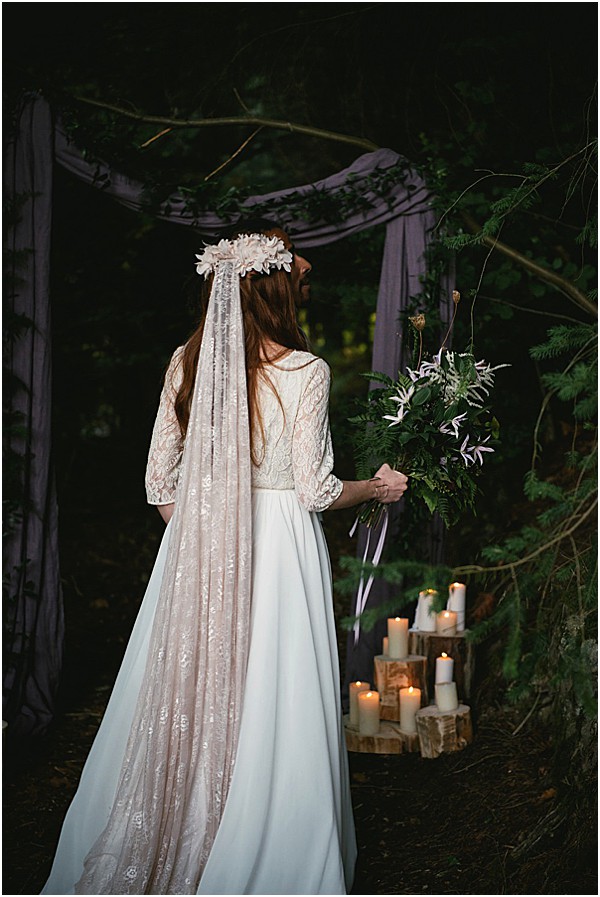 A bride stands with her back to the camera in an outdoor woodland ceremony setting, facing a rustic altar draped with muted mauve-grey fabric tied to tree branches. She wears a two-piece bridal outfit with a lace three-quarter-sleeve top and a flowing ivory skirt, paired with a long lace-edged veil and a white floral crown. She holds a loose, garden-style bouquet of dark green ferns, foliage, and small pale lavender blooms with trailing grey ribbons. To her right, white pillar candles of varying heights are arranged on raw wood log slices as decor, adding warm candlelight to the dark, moody forest surroundings. The overall styling theme is boho-woodland with a dark, moody atmosphere. Portrait shot from behind.