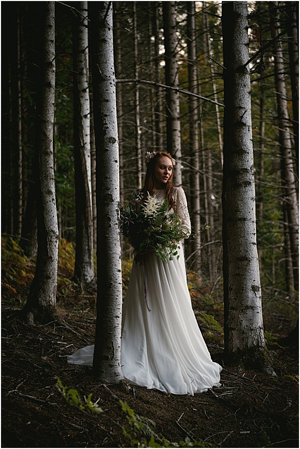 A bridal portrait taken outdoors in a dense forest setting with tall, slender birch-like trees and dark, moody undergrowth. The bride stands alone among the trees wearing a flowing ivory gown with long lace sleeves and a chiffon skirt, holding a loosely arranged bouquet composed of ferns, dark foliage, white thistle-like blooms, and trailing greenery in a predominantly green and deep plum palette. She wears a delicate floral hair accessory and her auburn hair is worn loosely down. The overall styling is boho-woodland with a dark, atmospheric tone created by low natural light filtering through the forest canopy. The shot is a full-length portrait with a wide enough frame to show the forest environment surrounding the subject.