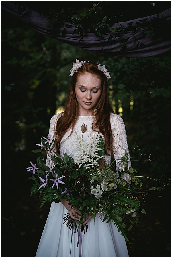 A bridal portrait taken outdoors in a wooded setting with dark, moody lighting. The bride has long red hair and wears a white lace long-sleeve dress with a flowy skirt, a delicate floral crown of small pale pink blooms, and a layered pendant necklace. She holds an oversized cascading bouquet composed of ferns, white astilbe, lavender star-shaped flowers, dark berries, small white wildflowers, and trailing greenery, tied with a lavender ribbon. A length of dusky purple fabric is draped overhead, framing the shot. The overall styling is bohemian with a dark, woodland aesthetic. Close-up portrait composition with the bride's gaze directed downward.