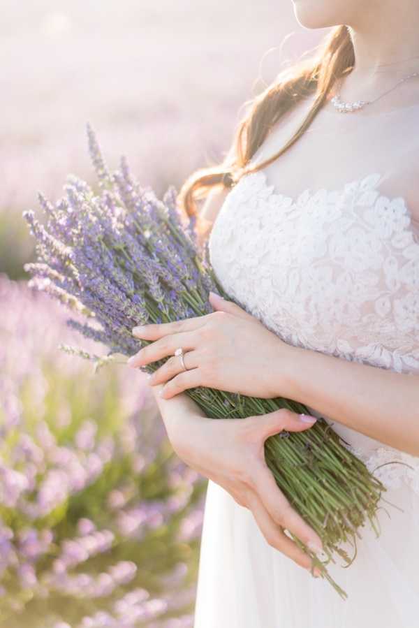 Close-up portrait of a bride standing outdoors in a lavender field, holding a simple bouquet of fresh-cut lavender stems tied together. The bride is wearing a white lace bodice dress with floral appliqué detailing and a delicate crystal necklace. Her engagement ring and wedding band are visible on her left hand as she cradles the bouquet. The image is softly lit with warm backlighting, creating a bright, airy feel, with the purple lavender field blurred in the background.
