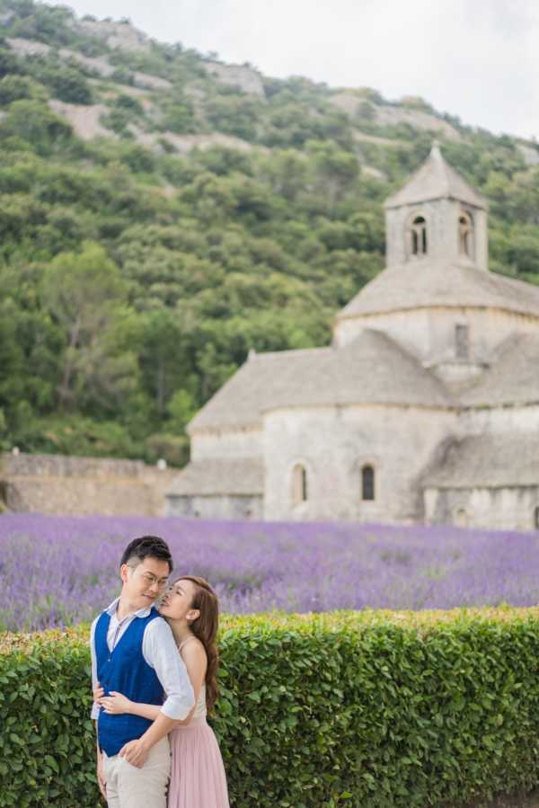 A couple poses outdoors in front of the Abbaye de Sénanque in Provence, with rows of purple lavender fields and a wooded hillside visible in the background. The man wears a blue vest over a light blue shirt with beige trousers, and the woman wears a strapless light pink skirt and nude top, resting her head against his shoulder as they embrace. The shot is a mid-length portrait with the historic Romanesque stone abbey softly blurred in the background. The lavender field provides a vivid purple color backdrop between the clipped green hedge in the foreground and the abbey beyond.