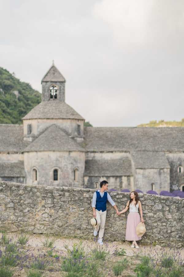 An engagement or pre-wedding couple portrait shot outdoors in front of the Abbaye de Sénanque, a Romanesque stone abbey in Provence, France. The couple stands holding hands against a low stone wall, with rows of lavender visible to the right in the background. The man wears khaki trousers, a light blue shirt, and a navy vest, holding a straw hat; the woman wears a blush pink midi skirt and a white sleeveless top, also holding a straw hat. The styling is casual and relaxed with a soft Provençal aesthetic. This is a full-length portrait shot with the abbey's bell tower and apse prominently featured in the background. Potential venue feature image.