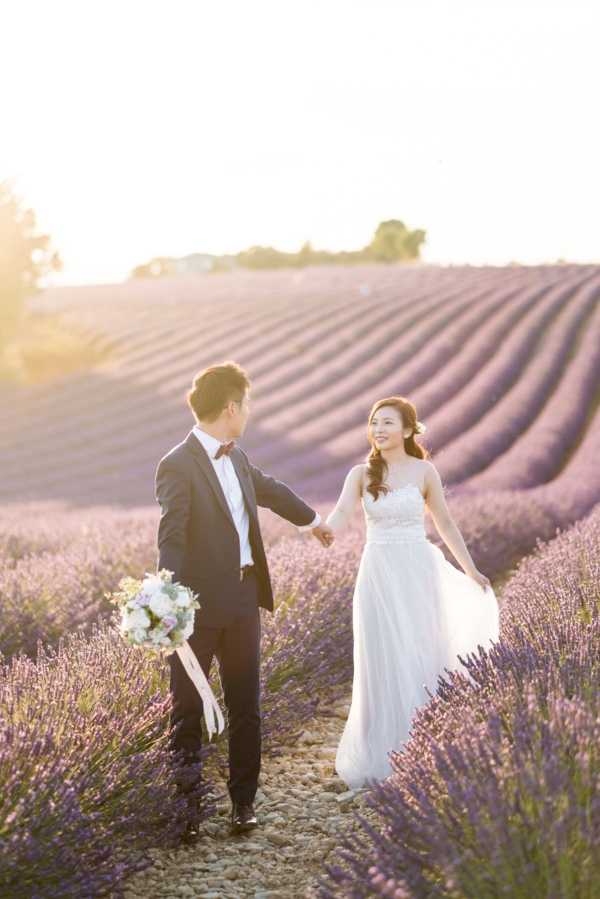A couple poses for a portrait session in a Provence lavender field, walking hand-in-hand along a path between the rows of purple lavender in bloom. The bride wears a floor-length white gown with a lace bodice and flowing skirt, while the groom is dressed in a dark navy suit with a black bow tie and white dress shirt. The groom carries a round bouquet of white and pale pink blooms with trailing white ribbons. The shot is taken at golden hour, with warm backlight creating a soft glow across the scene, and is framed as a mid-distance portrait capturing both figures full-length amid the lavender rows.