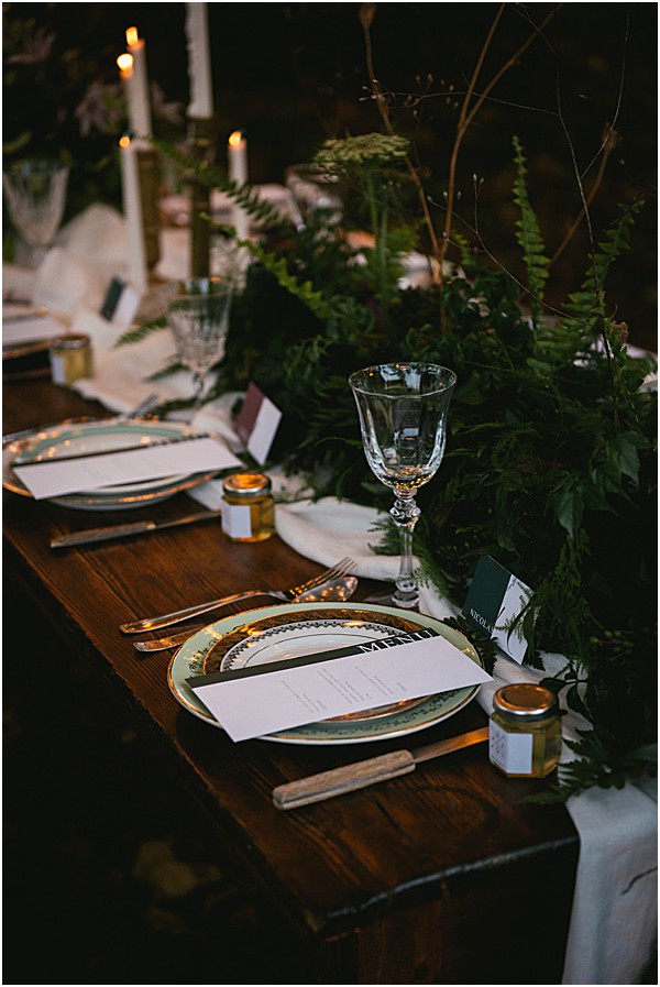 A close-up detail shot of a wedding reception tablescape set on a dark wood farm table. The place settings feature layered plates with gold-rimmed chargers, crystal glassware, and silver cutlery, with printed menu cards and dark green place cards. A dense greenery runner made of ferns and foliage runs along the center of the table, accompanied by tall white taper candles in gold candlestick holders providing warm ambient lighting. Small glass jars of honey are placed as favor items at each setting, and a white linen runner underlies the greenery, reflecting a dark, moody botanical styling theme.
