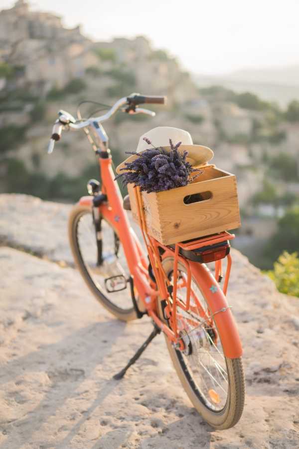 A styled detail shot featuring a coral-orange vintage-style bicycle parked on a rocky surface, with a wooden crate attached to the rear rack holding a bundle of purple dried lavender and a natural straw hat. The prop styling follows a Provençal rustic theme, with the lavender and wooden crate as key decorative elements. The background shows a blurred hilltop village typical of the South of France, suggesting an outdoor Provence-region setting. This appears to be a styled shoot prop or wedding decor detail image rather than a moment from the wedding day itself.