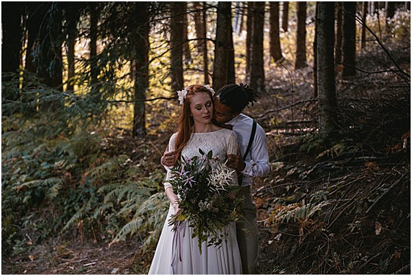 A couple portrait taken outdoors in a dense forest setting with tall pine trees and ferns in the background. The bride, with long red hair adorned with small white florals, wears a long-sleeve ivory lace-top gown and holds a large, loosely structured bouquet featuring deep purple thistles, greenery, and wild fern fronds in a boho style. The groom, dressed in a light grey shirt with dark suspenders, stands behind her with his face close to her cheek in an intimate pose. The composition is a mid-length portrait shot with warm, dappled natural light filtering through the trees.