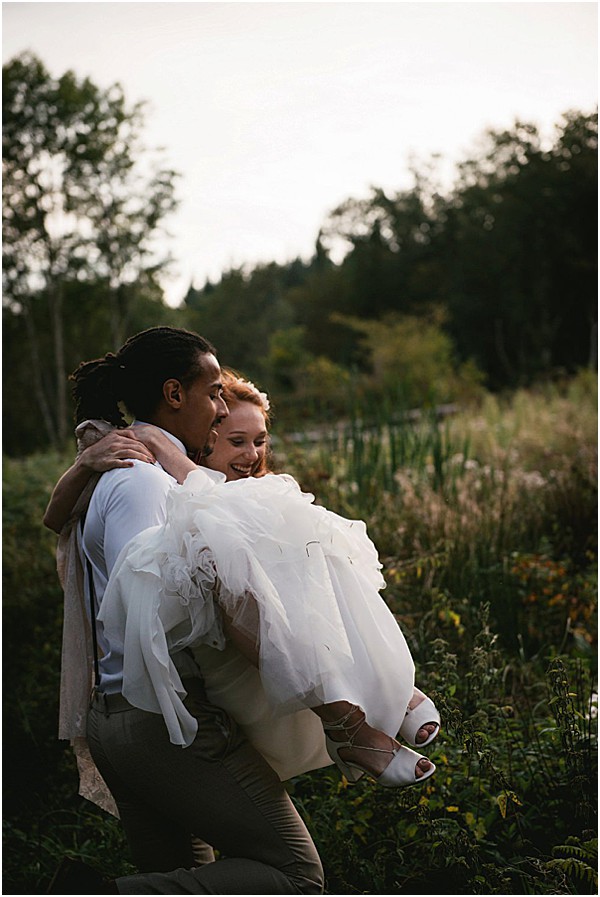 A couple portrait taken outdoors in a natural garden or wetland setting at dusk, with soft, fading light. The groom, wearing khaki trousers and tan suspenders over a light blue shirt, is carrying the bride in his arms while crouching slightly; she laughs and wraps her arms around his neck. The bride wears a layered white tulle dress and white open-toe heeled sandals, with short red hair. The composition is a medium portrait shot with the couple centered against a backdrop of dense greenery and tall grasses.