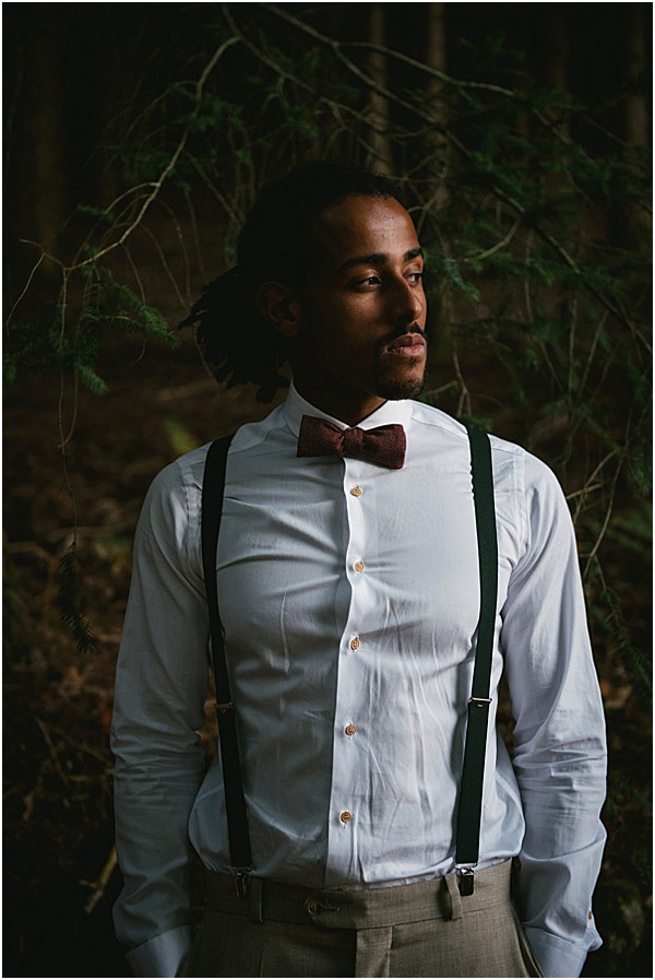 A portrait of a groom standing outdoors in a wooded setting with dark, moody ambient light. He is wearing a white dress shirt with gold buttons, dark green suspenders, and a burgundy bow tie, paired with light grey trousers. He has dreadlocks pulled back and a short beard, and his gaze is directed off-camera to one side. The composition is a close-up portrait shot with a shallow depth of field, keeping the forest background dark and out of focus, creating strong contrast against the white shirt.