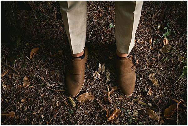 Close-up detail shot of a groom's feet, showing tan suede double monk-strap shoes paired with light beige linen trousers. The shot is taken from above, looking down at the shoes against a forest floor covered in fallen leaves and pine needles. The dark, moody lighting and rustic outdoor woodland setting suggest a natural or boho styling approach.