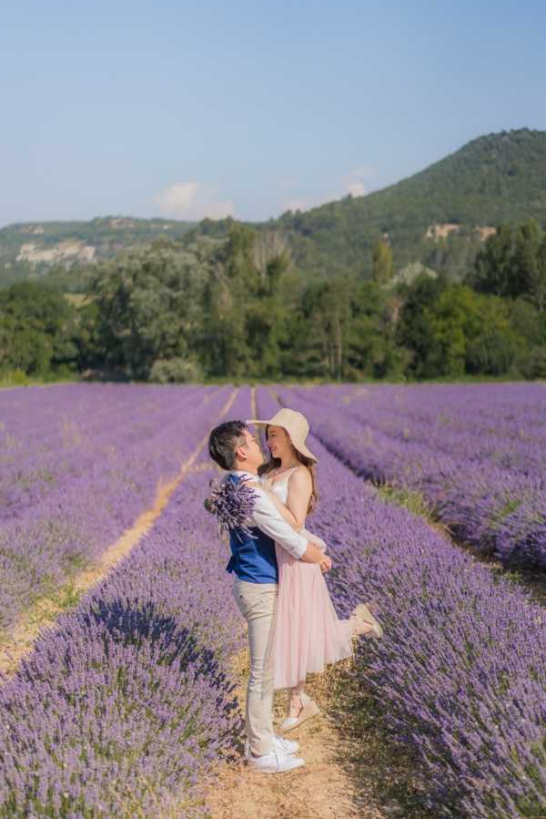 A couple poses together in an outdoor engagement or pre-wedding portrait session set in a Provençal lavender field in full purple bloom, with rows of lavender extending into the background. The woman wears a light blush pink midi skirt, a white sleeveless top, white sneakers, and a wide-brim straw hat, and holds a small bunch of freshly cut lavender; the man wears a navy blue vest over a white shirt with beige trousers. The two face each other closely in an embrace, with one of her feet lifted off the ground. The shot is a medium full-body portrait framed along the central path between lavender rows.