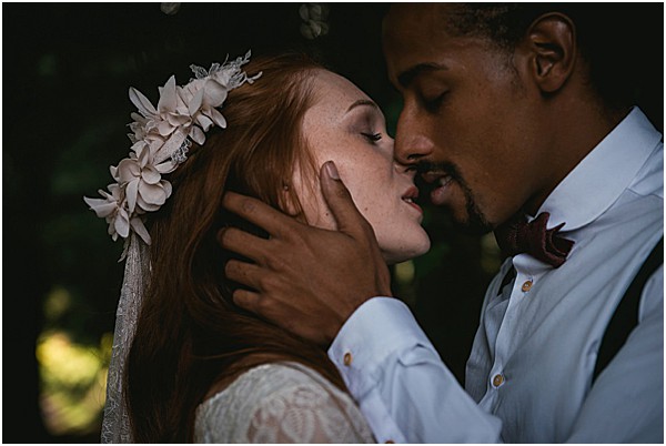 Close-up portrait of a bride and groom sharing a kiss outdoors against a dark, blurred green foliage background. The bride has long red hair and wears a floral headpiece made of cream fabric flowers with a veil, along with a lace dress. The groom wears a light grey suit jacket with a deep burgundy bow tie, and his hand gently cups the bride's face. The styling has a boho-romantic feel, with the floral headpiece and lace detailing as intentional design choices. The image has a moody, low-light quality with dark tones surrounding the couple.