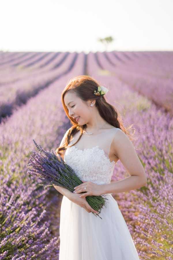 A bridal portrait taken outdoors in a Provence lavender field, with rows of purple lavender extending into the background. The bride wears a white gown with a lace appliqué bodice and flowing chiffon skirt, paired with a pearl necklace and drop earrings. She holds a hand-tied bouquet of fresh-cut lavender stems and has a small white flower tucked into her half-up, loose dark hair. The shot is a mid-length portrait with soft natural light and a shallow depth of field that blurs the lavender rows behind her.