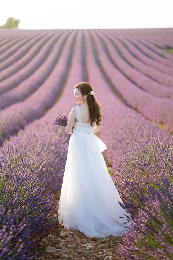 A bride stands alone in an outdoor lavender field, shot from behind at a three-quarter angle as she turns slightly to her left. She wears a white strapless ball gown with a lace-embellished bodice, a peplum detail at the waist, and a full tulle skirt, and holds a small bouquet of fresh lavender sprigs. Her long dark hair is worn down with a white flower hair accessory. The rows of purple lavender extend symmetrically into the background, creating strong leading lines, and the image is lit with warm golden-hour light. This is a medium portrait shot taken at ground level during what appears to be a bridal portrait or pre-wedding session in Provence.