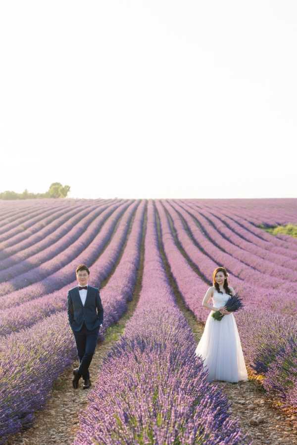 A couple poses for a portrait in an outdoor lavender field in Provence, with long rows of purple lavender extending to the horizon under bright, flat natural light. The groom stands on the left wearing a navy suit with a burgundy bow tie, while the bride stands on the right in a strapless white ballgown with a full tulle skirt, holding a bouquet of bundled dried or fresh lavender stems. The two are positioned several feet apart, each standing between rows of lavender, facing the camera. The wide shot composition uses the converging lines of the lavender rows as a leading element through the frame.