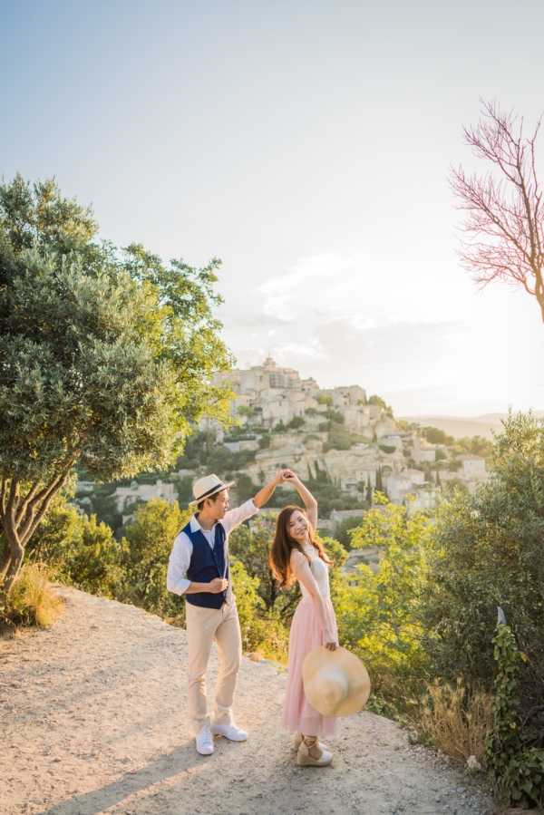 A couple poses for an outdoor portrait session on a dirt path overlooking a hilltop Provençal village, likely Gordes in the Luberon region of southern France. The man wears a navy vest over a white shirt with beige trousers and a straw hat, while the woman wears a light blush pink midi dress with espadrille wedges and holds a large wide-brim straw hat; he spins her by one hand as they both laugh. The shot is taken during golden hour, bathing the scene in warm amber light. This is a full-length couple portrait with the historic perched village and surrounding vegetation visible in the background.