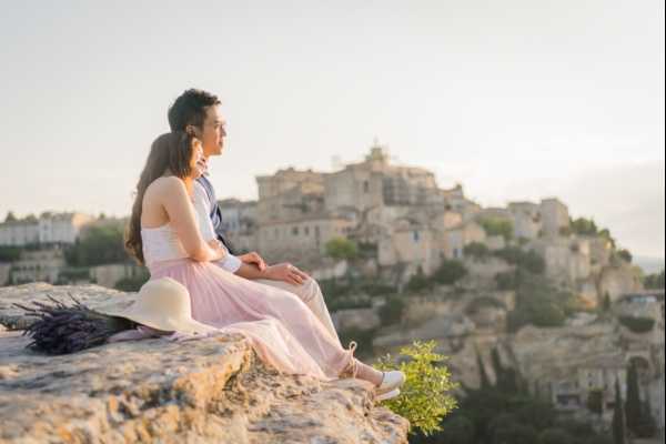 A couple sits together on a rocky ledge overlooking the hilltop village of Gordes in Provence, both gazing into the distance in a relaxed, candid pose. The bride wears a white off-the-shoulder top with a blush pink tulle skirt and white heeled sandals, while the groom is dressed in a light grey suit. A white sun hat and a small bunch of dried lavender rest on the rock beside them. The shot is a medium-wide portrait taken at golden hour, with warm soft light across the scene.