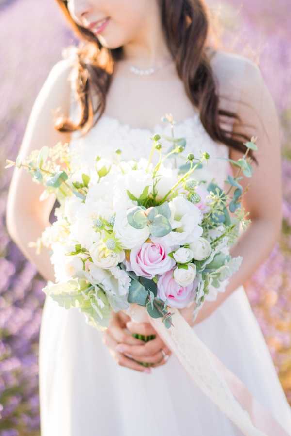Close-up portrait of a bride holding her bouquet outdoors, shot from the chin down with the face partially cropped at the top of the frame. The bride wears a white sleeveless dress with lace or floral detailing at the neckline and a delicate pearl necklace. The bouquet features white peonies, white and blush ranunculus, soft pink roses, scabiosa buds, and eucalyptus foliage, tied with a trailing ivory ribbon. The background is softly blurred lavender, consistent with a lavender field setting, creating a light purple bokeh effect. The overall styling is romantic and garden-inspired with a soft pastel palette of white, blush pink, and sage green.