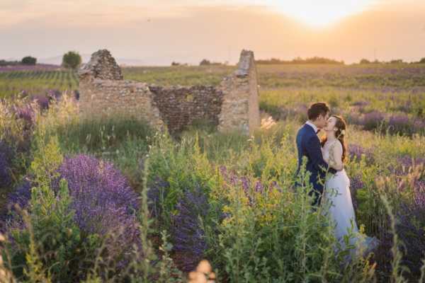 A couple shares a kiss during a portrait session in a lavender field at golden hour, with warm orange and pink tones from the setting sun illuminating the scene. The bride wears a white gown with a fitted lace bodice, and the groom is dressed in a navy blue suit. They are surrounded by purple lavender bushes and yellow wildflowers, with the ruins of a small stone structure visible behind them and rows of a vineyard or crop field stretching into the background. The shot is a mid-range portrait framed within the natural vegetation, capturing the couple slightly off-center.