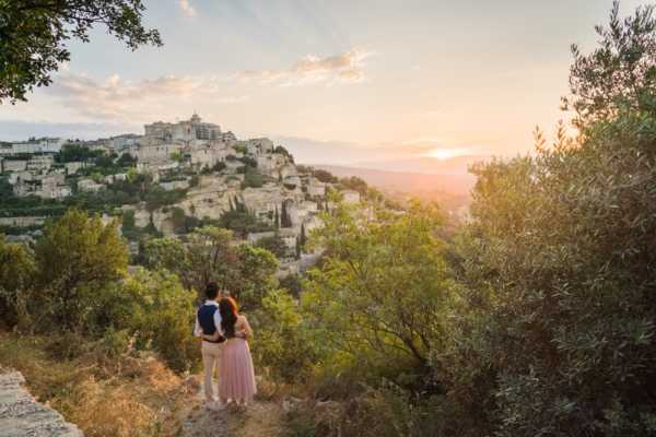 A couple stands together on an elevated hillside viewpoint, looking out over a historic perched village — identifiable as Gordes in Provence — during golden hour at sunset. The groom wears dark trousers and a navy vest, while the bride wears a flowing blush-pink gown. The wide-angle shot frames the couple from behind in the lower center, surrounded by olive trees and Mediterranean vegetation, with the stone village buildings cascading down the rocky hillside in the background and warm sunset light flooding the horizon. This is an outdoor portrait session in a natural, unposed style typical of engagement or post-ceremony couple photography.