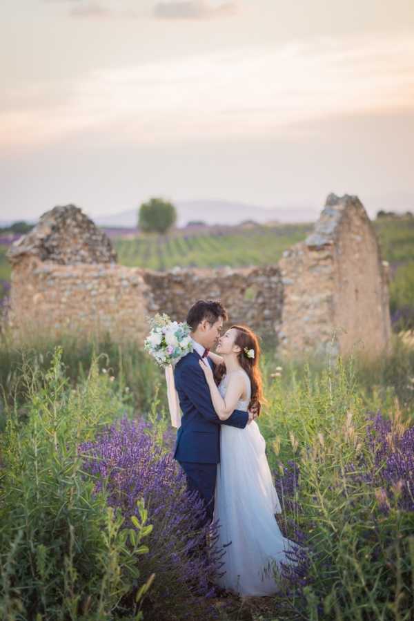 A couple poses in an outdoor portrait session amid a flowering lavender field in Provence, with the remains of a ruined stone structure visible in the background and rows of lavender crops stretching into the distance. The groom wears a navy blue suit with a burgundy bow tie, and the bride wears a light blue-grey tulle gown with a white floral hair accessory. The bride holds a loose bouquet of white and blush blooms with trailing ribbons. The shot is a medium portrait framed from the knees up, with the couple facing each other closely in an about-to-kiss moment during golden hour light.