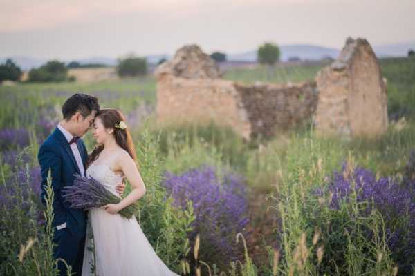 A couple poses for a portrait in an outdoor lavender field at dusk, with a crumbling stone ruin visible in the background. The bride wears a strapless ivory ball gown and carries a loose bouquet of fresh lavender stems, with a small white flower accessory in her hair; the groom wears a navy suit with a burgundy bow tie. The two stand close together, foreheads nearly touching, surrounded by tall purple lavender plants. The shot is a mid-range portrait with soft, warm evening light and a slightly hazy backdrop.