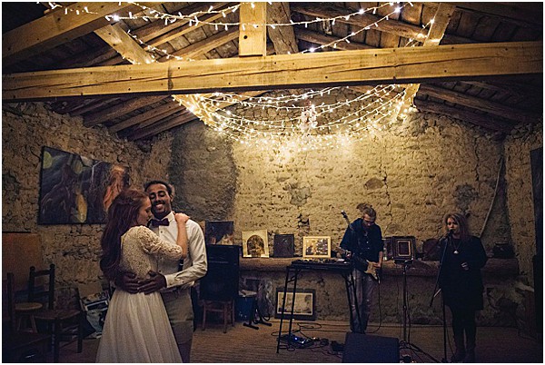 A couple shares their first dance inside a rustic stone barn venue during the reception, with the bride wearing a long-sleeved lace white gown and the groom in a white shirt with a dark bow tie. The barn has exposed wooden beam rafters strung with warm white fairy lights that create a canopy effect across the ceiling. In the background, two musicians perform at a makeshift stage setup against the stone wall, with what appear to be framed artworks and equipment arranged on a wooden table. The wide shot captures the warm, intimate atmosphere of the dimly lit interior with a rustic, boho styling theme.