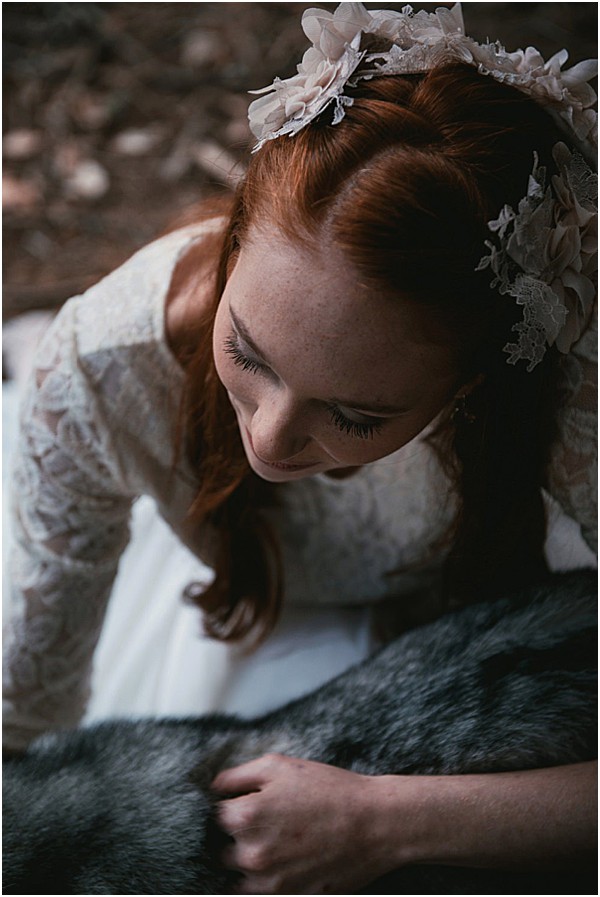 A close-up portrait of a bride with red hair, shot from slightly above. She is looking downward and smiling, wearing a long-sleeve ivory lace dress and an ivory floral headpiece made of fabric flowers and lace detailing. She is leaning over or resting on a dark grey faux fur throw or animal skin. The setting appears to be outdoors in a wooded area. The styling has a boho-romantic feel, with natural, undone hair and a delicate floral crown. The image has a moody, low-contrast edit with muted, cool-toned shadows.