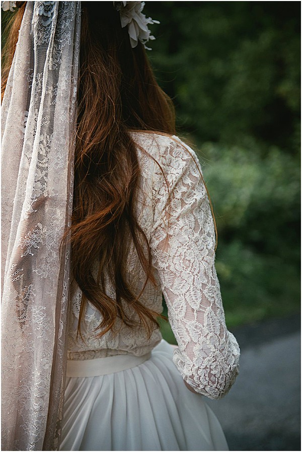 A close-up detail portrait of a bride shot from behind, focusing on the back of her ivory lace long-sleeve crop top paired with a flowing white chiffon skirt. Her long auburn wavy hair falls over the lace, and she wears a white floral crown with small white blooms. A lace-edged veil drapes over her left shoulder. The styling is boho-romantic with the two-piece separates, floral crown, and delicate veil detailing. The composition is a tight portrait crop emphasizing the texture of the lace against the bride's hair.