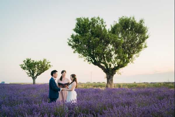 An intimate outdoor ceremony taking place in a lavender field in full purple bloom, likely in Provence. Three people are present — a groom in a navy suit, a bride in a white gown with her hair up, and an officiant in a blush/light pink dress holding an open book and reading the ceremony. In the background, a naturally heart-shaped tree provides a striking focal point against a soft pastel evening sky. The setting is rustic and natural with no visible decor beyond the lavender surroundings. Wide portrait shot capturing all three figures at mid-distance within the expansive field.
