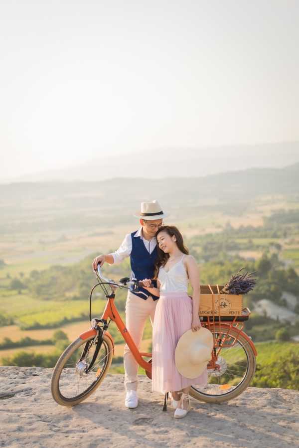 A couple poses together outdoors on an elevated rocky overlook with a sweeping valley landscape visible in the background, shot as a full-length portrait. The man wears a navy blue vest over a white shirt with beige trousers and a straw fedora hat, while the woman wears a white sleeveless top with a blush pink midi skirt and holds a wide-brim straw hat. They stand beside an orange vintage-style bicycle fitted with a wooden crate rear basket containing a bunch of dried lavender, with a 'Location Vente' label visible on the crate. The styling is relaxed and Provençal-inspired, with warm golden-hour light casting a soft glow across the scene.