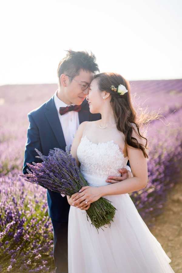 A couple poses together in a portrait shot taken outdoors in a blooming lavender field, likely in Provence. The bride wears a white lace-bodice A-line gown with a tulle skirt, a pearl necklace, and a white flower hair accessory, and carries a hand-tied bouquet of fresh purple lavender stems. The groom wears a navy suit with a white dress shirt and a deep burgundy bow tie. The two face each other closely in an intimate pose, with the groom's arms around the bride. The background is filled with rows of purple lavender in full bloom, rendered soft and out of focus, with bright backlight creating a warm, hazy glow.