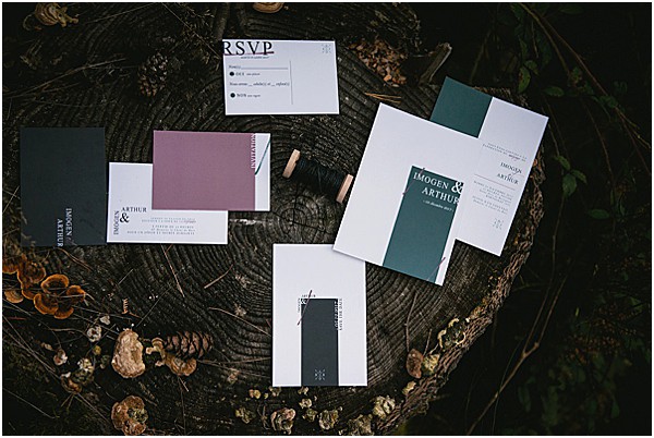 A flat lay detail shot of a wedding stationery suite for 'Imogen & Arthur' arranged on a large weathered tree stump. The suite includes invitation cards, an RSVP card, and additional inserts styled across two color stories: one featuring deep forest green color-block panels on white card stock, and another featuring muted mauve/dusty purple panels on white with dark charcoal text. A small wooden cylindrical wax seal or cord bobbin is visible among the pieces. The typography is modern and minimal, using clean serif and sans-serif fonts. The overall design aesthetic is contemporary and moody, complemented by the dark, textured wood surface and small natural fungi visible around the edges of the stump.