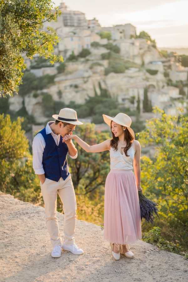 An outdoor couple portrait session set against the backdrop of a hilltop village, likely Gordes in Provence. The man wears cream chinos, a white shirt, a navy blue waistcoat, white sneakers, and a straw fedora with a dark band, and is bending to kiss the woman's hand. The woman wears a white sleeveless top, a blush pink tulle midi skirt, flat espadrille-style shoes, and a wide-brim straw sun hat, and holds a bundle of dried deep purple lavender. The composition is a full-length portrait shot in warm golden-hour light with the rocky, pale limestone village architecture visible and softly blurred in the background.