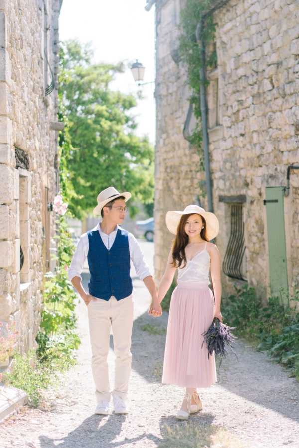 A couple walks hand-in-hand along a narrow gravel lane between old stone buildings in what appears to be a Provençal village, in a portrait-style engagement or pre-wedding shoot. The man wears a navy blue waistcoat over a white shirt with cream trousers and a straw fedora, while the woman wears a white spaghetti-strap top, a dusty pink midi tulle skirt, wedge sandals, and a wide-brim straw sun hat. She carries a small bunch of dried lavender. The styling has a relaxed, rustic French countryside feel, shot in bright natural daylight with soft shadows on the gravel path.