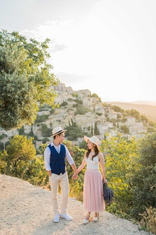 A couple poses outdoors on a rocky overlook with the perched hilltop village of Gordes in Provence visible in the background during golden hour. The man wears a navy blue vest over a white shirt with beige trousers, white sneakers, and a straw fedora, while the woman wears a blush pink tulle midi skirt, white sleeveless top, white lace-up flats, and a straw hat, holding a bunch of dried lavender. They are holding hands and looking at each other, styled in a relaxed, boho-casual aesthetic with a distinctly Provençal feel. The shot is a full-length portrait with warm golden light bathing the scene.