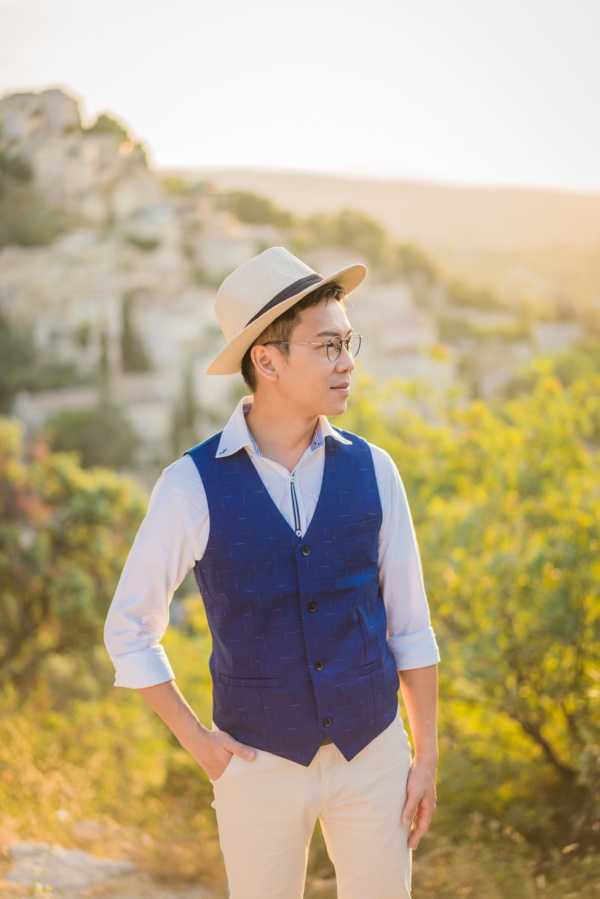 An outdoor portrait of a groom standing alone in warm golden-hour light, gazing to one side with his hands in his pockets. He is dressed in a navy blue patterned waistcoat over a white dress shirt with the sleeves rolled up, paired with cream trousers, round-frame glasses, and a cream fedora-style hat with a black band. The background features a rocky hilltop landscape with dense green shrubs, consistent with the Provence or Les Baux-de-Provence region of southern France. The shot is a medium portrait composition with a shallow depth of field, keeping the subject sharp against the softly blurred, sun-drenched landscape behind him.