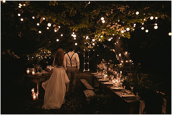The couple walks together through an outdoor nighttime reception set beneath a large tree draped with globe string lights. The bride wears a flowing white gown with a full skirt, and the groom is dressed in a white shirt with dark suspenders and light trousers. Long rustic wooden banquet tables are set on either side, lit by clusters of white taper candles and scattered tea lights at ground level, with greenery used as table runners. A wooden swing hangs from the tree branches above. The overall styling is boho-rustic with a warm, candlelit atmosphere created entirely by the globe lights and candles. Wide shot taken from behind the couple, emphasizing the moody, dark surroundings contrasted against the warm glow of the lighting.