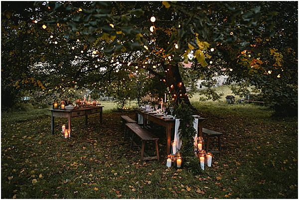 An outdoor reception tablescape set beneath a large tree at dusk, styled in a rustic, intimate boho aesthetic. A long dark wooden farm table with matching wooden benches is dressed with a white linen table runner, tall white taper candles, and what appears to be a greenery garland running along the center, with glassware and place settings for approximately 10–12 guests. Clusters of white pillar candles in varying heights are arranged on the ground around the base of the tree, and warm string lights are strung through the tree branches overhead, creating a soft amber glow against the dim evening light. A smaller wooden side table with additional candles and bottles is visible to the left, and the overall color palette is warm amber and deep green. Wide establishing shot capturing the full table setup and surrounding garden.