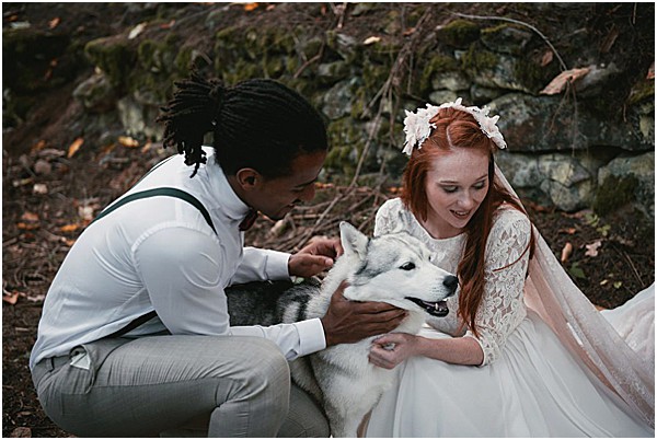 A couple crouches outdoors petting a Siberian Husky dog in a casual, candid portrait shot. The groom wears a white turtleneck top with grey trousers and green suspenders, while the bride wears a long-sleeved lace and tulle white gown with a white floral crown and veil in her red hair. Both are smiling and focused on the dog rather than the camera. The styling has a relaxed, boho-woodland feel, shot against a rocky backdrop with bare branches.