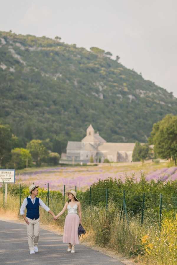 A couple walks hand-in-hand along a road in an outdoor portrait shot, with the Abbaye de Sénanque visible in the background surrounded by purple lavender fields in Provence. The groom wears beige trousers, a white shirt, and a navy vest with a straw hat, while the bride wears a white strappy top, a blush pink tulle midi skirt, white heels, and a straw hat, and carries a small bunch of fresh lavender. The styling is relaxed and romantic with a Provençal countryside feel. The composition is a full-length portrait taken from a medium distance, showing both figures in motion against the scenic backdrop.