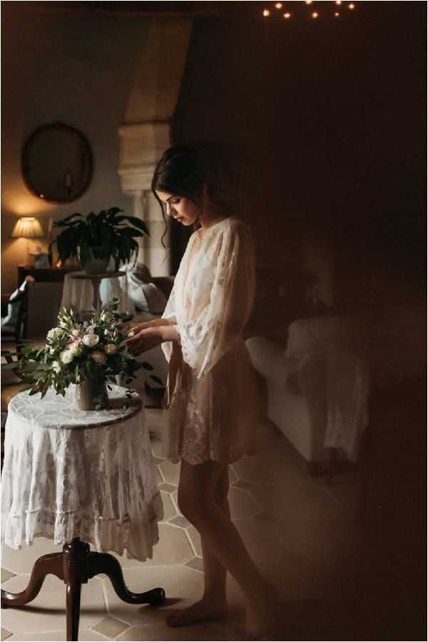 A bride is photographed getting ready in a dimly lit interior room, standing barefoot beside a small antique side table draped with a white lace tablecloth. She is wearing a sheer white lace robe and is looking down, touching a bouquet of blush pink roses, white flowers, and greenery arranged in a ceramic vase. The room features warm ambient lighting from a table lamp, fairy lights visible in the upper background, a round wall mirror, and a large leafy houseplant. The overall palette is warm and dark with moody shadows. The composition is a medium full-length portrait shot with a romantic, boho styling aesthetic.