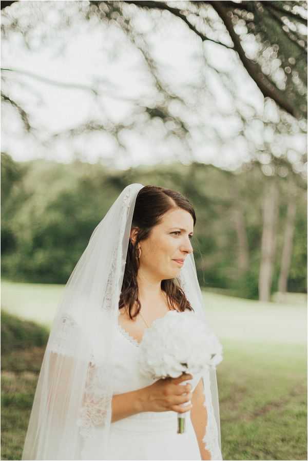 A bridal portrait taken outdoors in a garden or parklike setting, with trees and open lawn visible in the soft-focus background. The bride, a dark-haired woman, wears a white lace-sleeved gown and a long cathedral-length veil with a lace trim edge, and holds a compact bouquet of white peonies. She is gazing slightly off to one side with a natural expression. The shot is a medium portrait composition with shallow depth of field, captured in soft, diffused natural light.