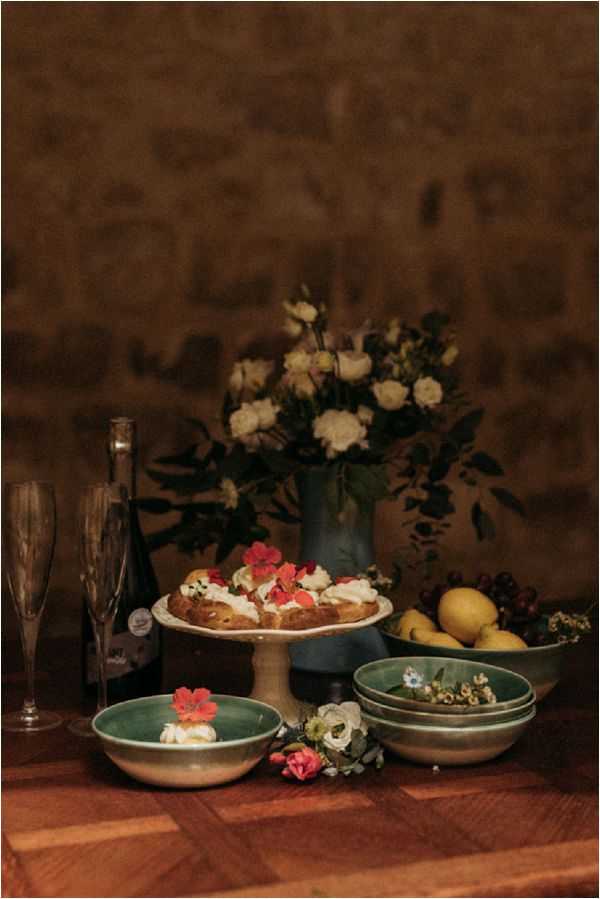 A close-up styled tablescape shot featuring a rustic dessert display set on a warm-toned wooden surface against an exposed stone wall backdrop. A ceramic pedestal cake stand holds a pastry topped with cream and red edible flowers, flanked by stacked green ceramic bowls, a bowl with cream and a red flower garnish, a small bowl of lemons and berries, and loose scattered florals including red and white blooms. A blue ceramic vase holds an arrangement of ivory roses, white lisianthus, and dark foliage in the background, alongside a dark wine bottle and a smoked grey champagne flute. The overall styling palette combines deep teal, terracotta red, ivory, and warm wood tones with a moody, intimate rustic aesthetic. The image is a vertical portrait-orientation detail shot taken in low, warm ambient light.