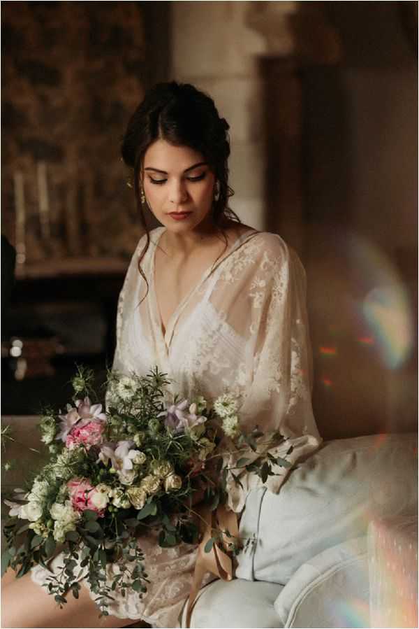 A bride sits indoors on a light grey upholstered seat, looking downward with a calm expression. She wears a sheer ivory lace robe or cover-up with long sleeves and holds a large, loosely arranged bridal bouquet featuring pink peonies, cream garden roses, white scabiosa, soft lavender clematis, and abundant eucalyptus and greenery with trailing champagne ribbons. Her dark hair is styled in a loose updo with soft face-framing pieces, and she wears small gold earrings. The background suggests a stone interior space with warm, dim ambient light, and a visible lens flare with prismatic colors appears on the right side of the frame. The shot is a close-to-medium portrait with a shallow depth of field, styled in a romantic, organic, boho-inflected aesthetic.