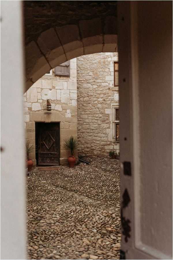 A venue detail shot framed through a partially open white door, looking through a stone archway into a cobblestone courtyard. The courtyard features an aged wooden door with decorative ironwork, flanked by terracotta pots with spiky green plants and a hanging lantern above the entrance. The building is constructed from cut limestone blocks, giving a historic French château or manor character. No people are present in the image. Potential venue feature image.