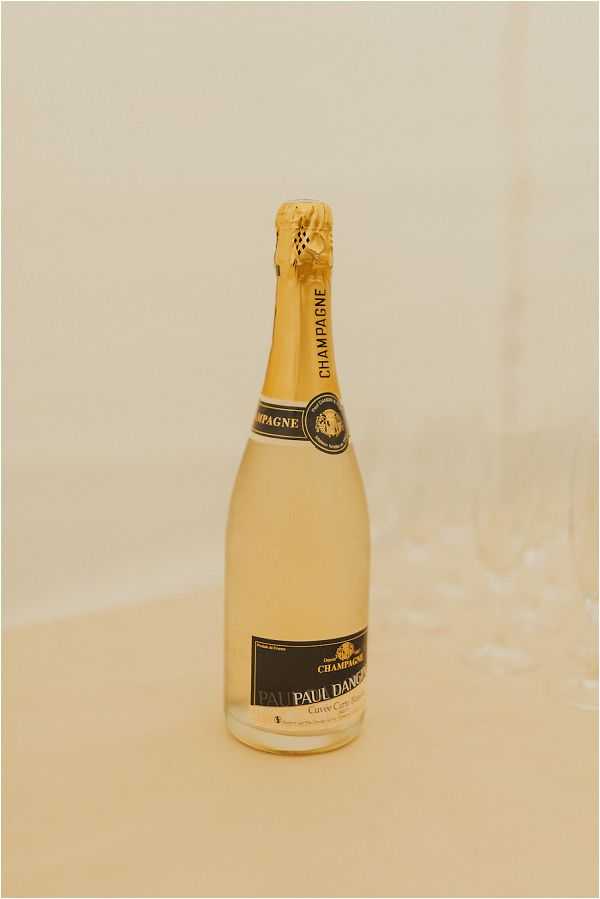 Close-up detail shot of a bottle of Paul Danco Champagne Cuvée Carte with gold foil neck and a black and gold label, placed on a cream-colored surface with what appears to be champagne flutes slightly out of focus in the background. The image has a warm, slightly overexposed tone with a cream and ivory palette throughout. No people are visible; this appears to be a reception or getting-ready detail photograph.